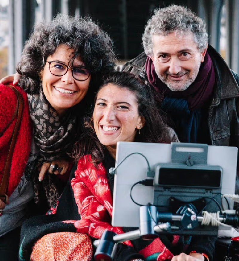 Photo of a young adult daughter using a wheelchair with an attached AAC device as she posing with her mother and father.