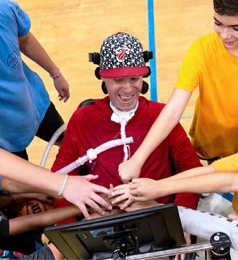 Photo of a basketball coach using a wheelchair with an attached AAC device as he's smiling and huddled up with his excited players in the middle of the basketball court.