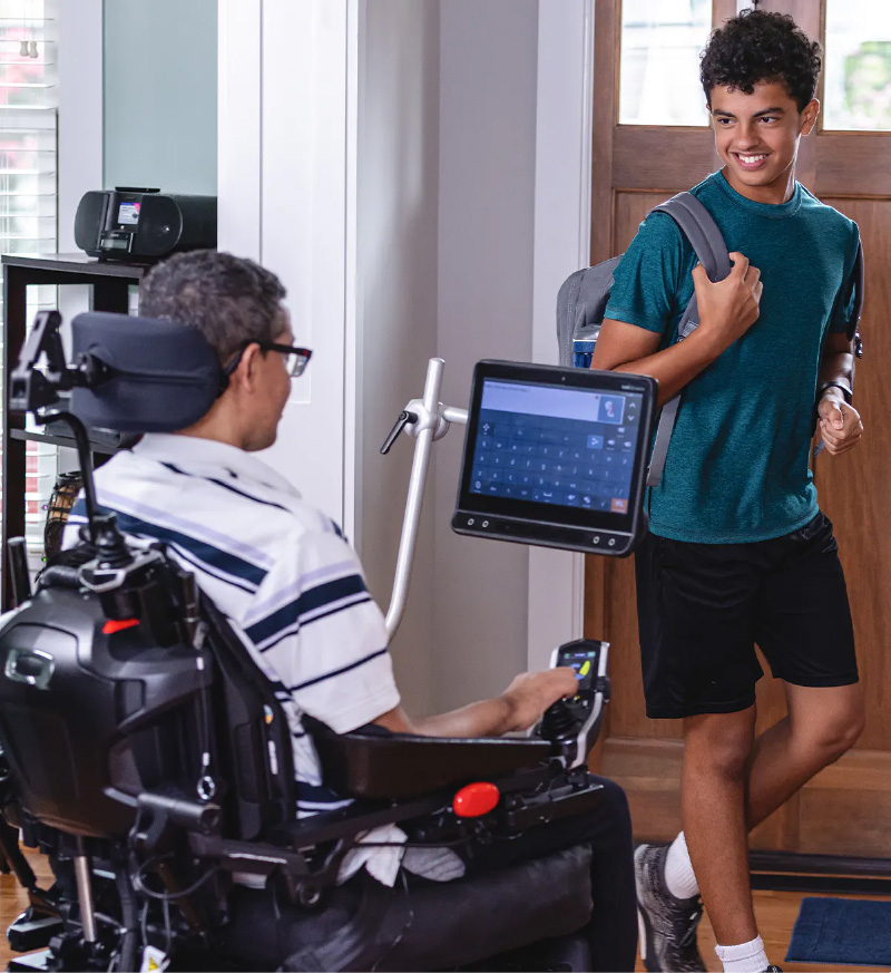 Photo of a father using a wheelchair with an attached AAC device greets his teenage son coming home with a backpack slung over his shoulder.