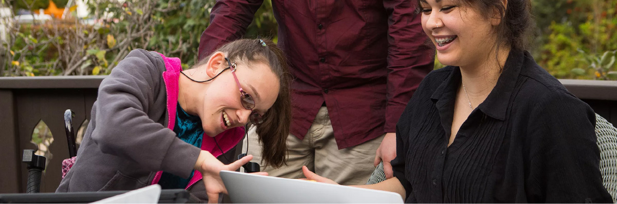 Photo of a young woman using a wheelchair with an AAC device next to her and communicating with another young woman at an outdoor table.