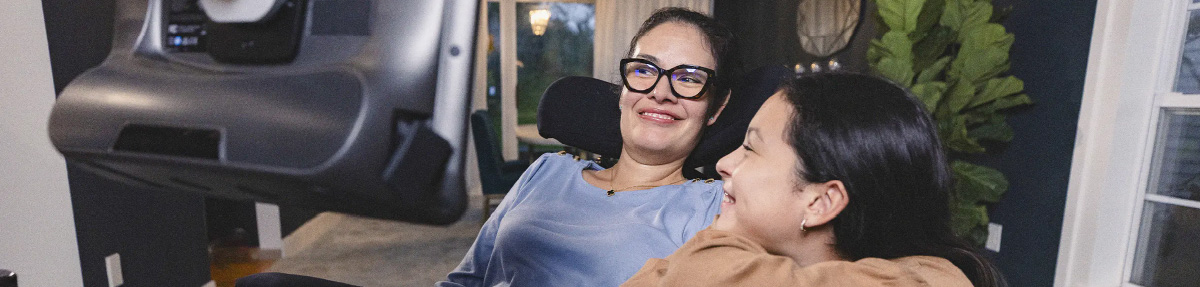 Photo of a mother using a wheelchair with an attached AAC device and having a conversation with her smiling teenage daughter.